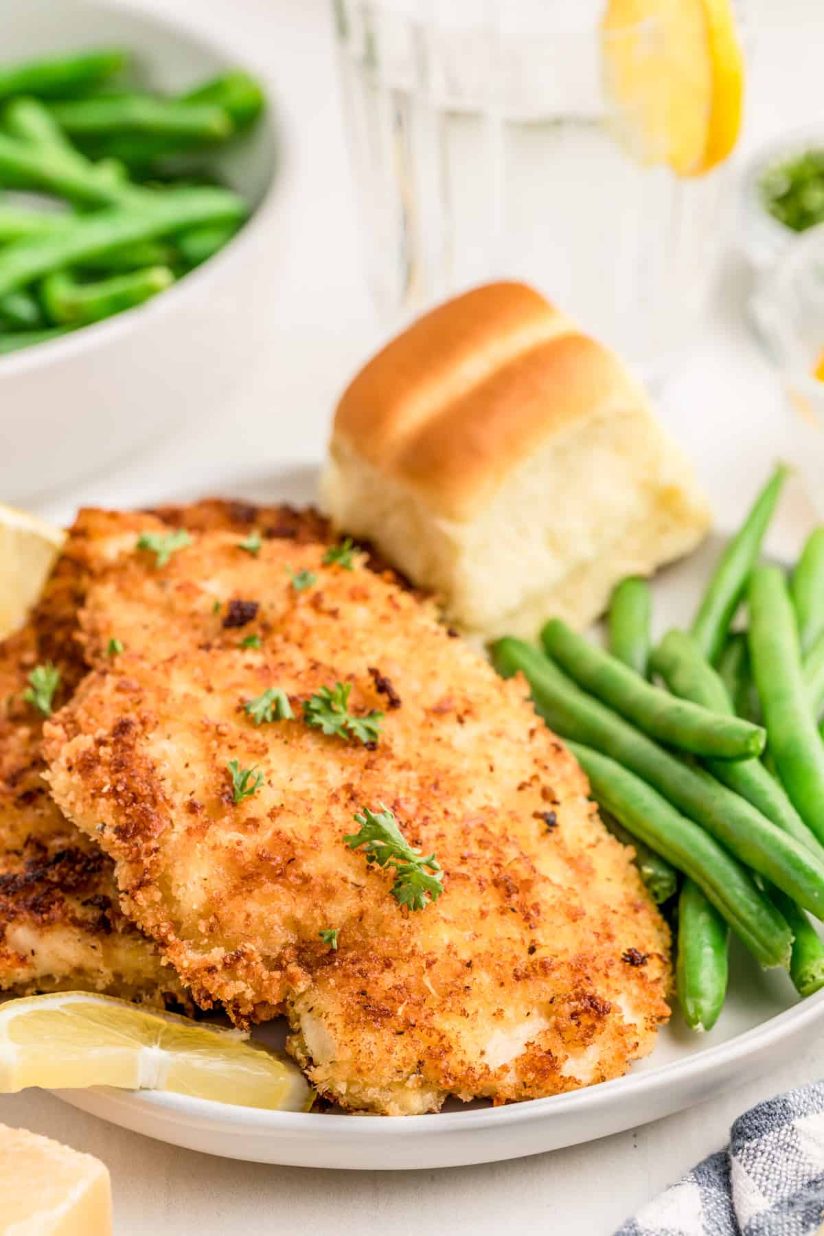 Parmesan Crusted Chicken on white plate with green beans, bread and lemon slice.
