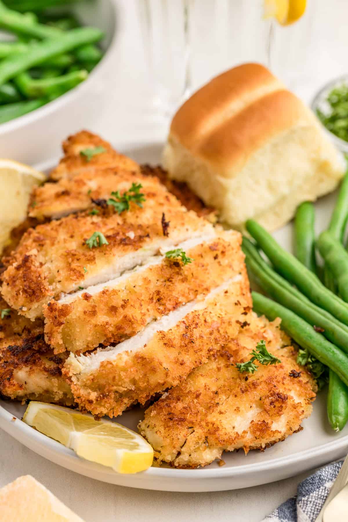 Cut up Parmesan Crusted Chicken on white plate with green beans, bread and lemon slice.