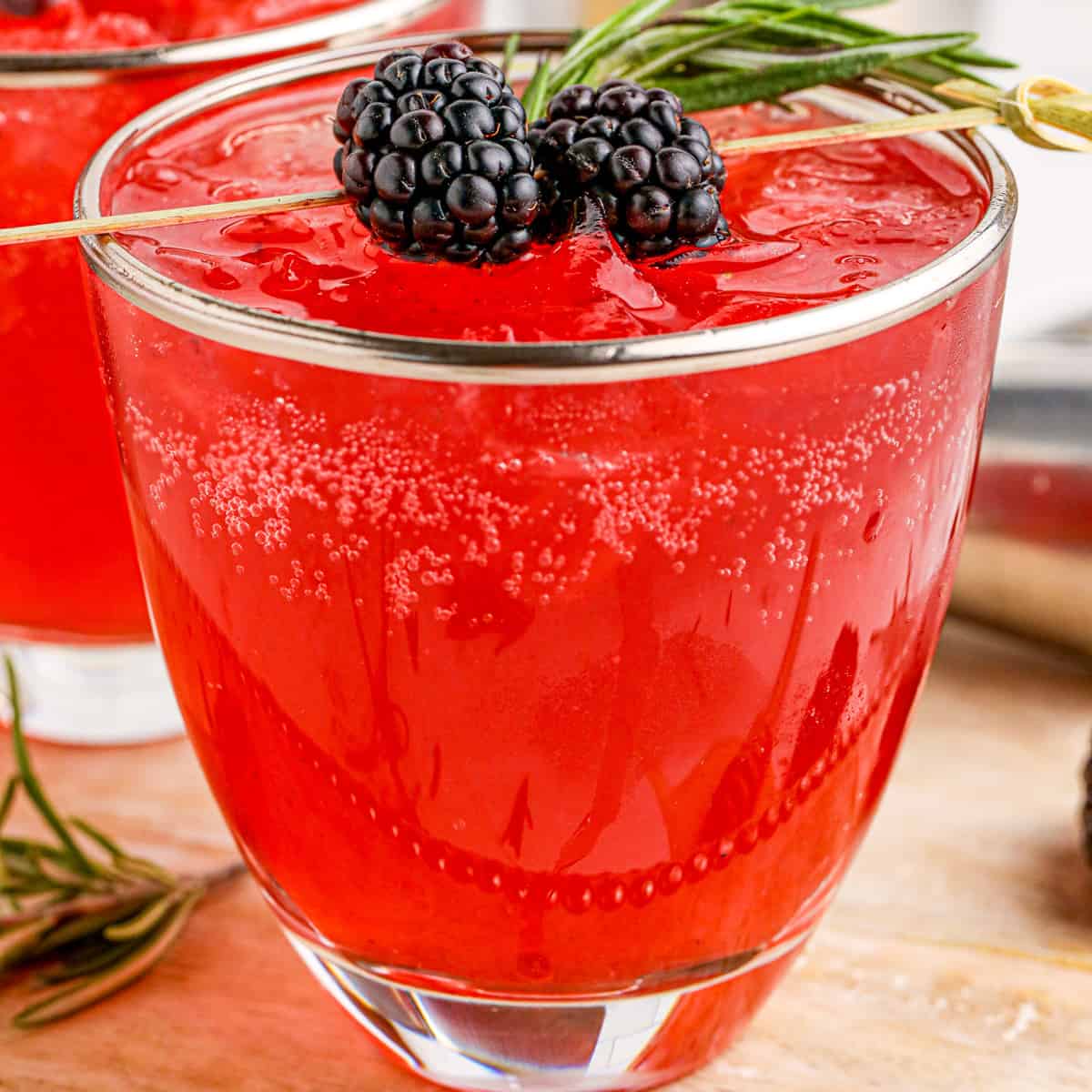 Close up square image of drink in metal rimmed glass garnished with blackberries and thyme.