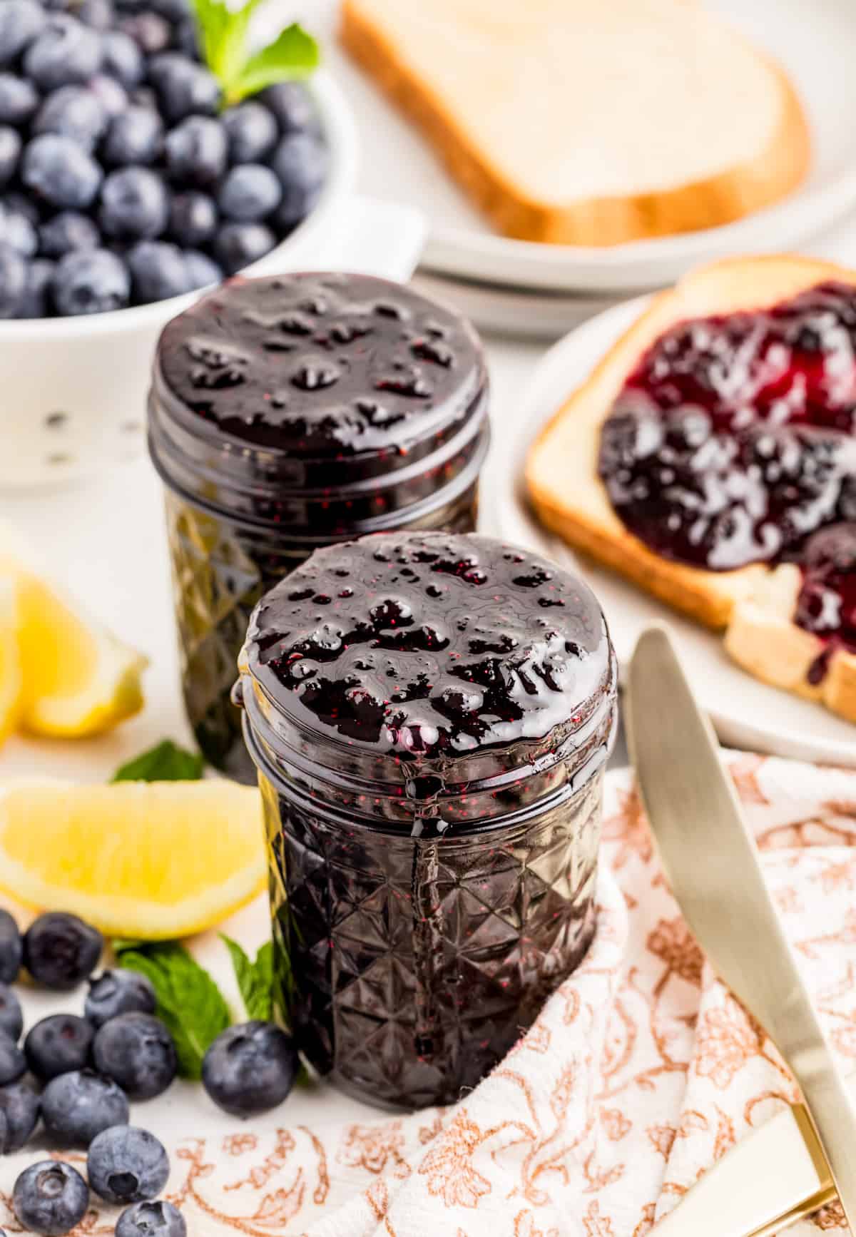 Two jars of jam with blueberries in background and jam spread on toast on the side of jars.