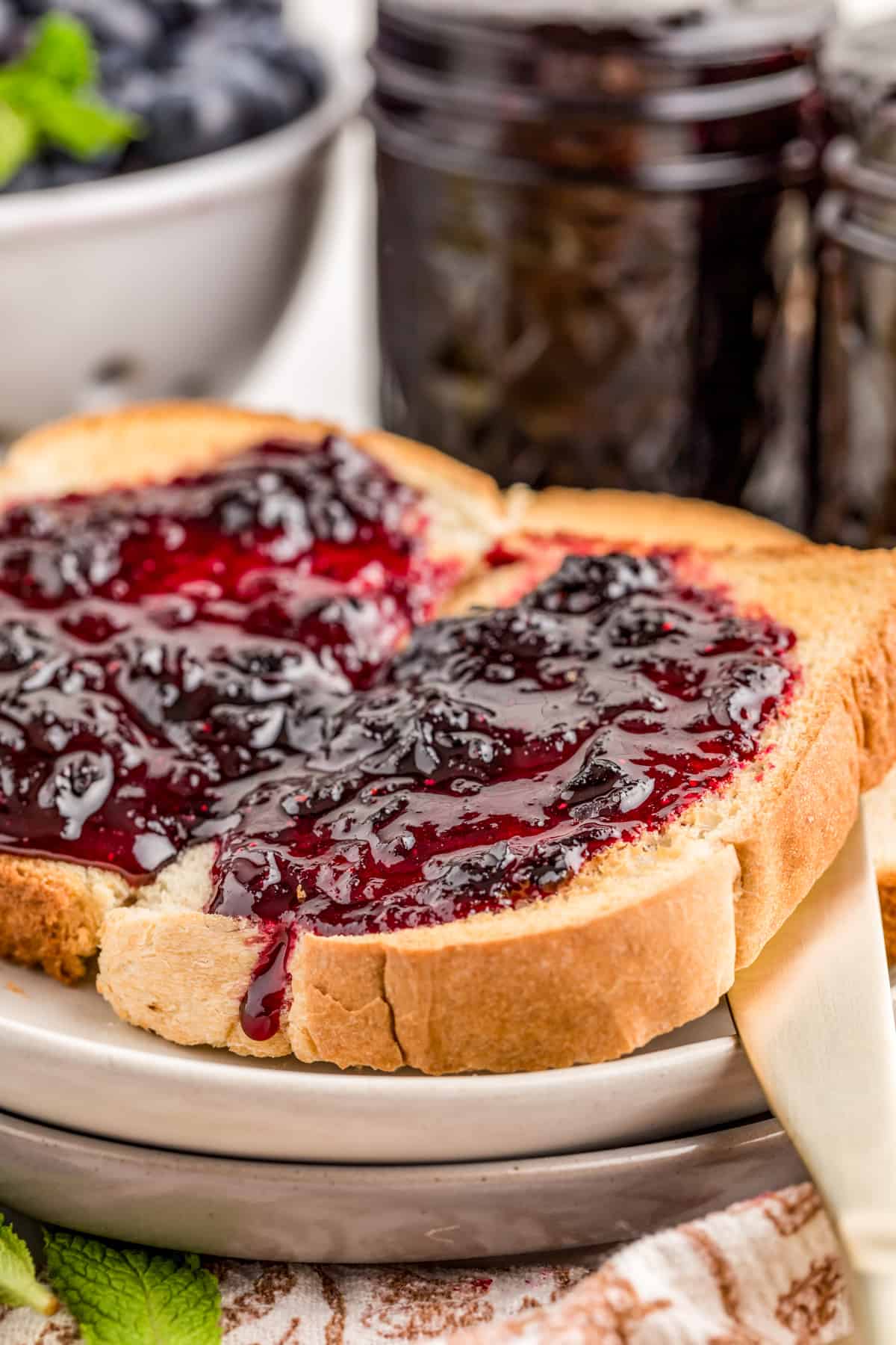 Jam spread on a cut piece of toast on white plate with jam in mason jar in background.