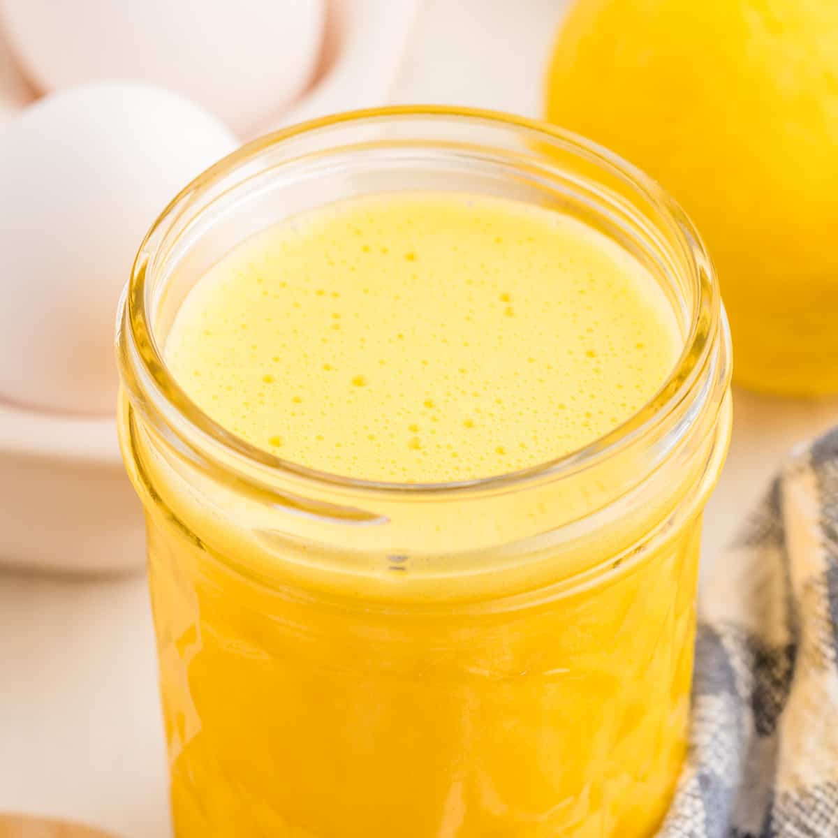 Close up square image of top of jar with sauce in it with eggs and lemon in background.