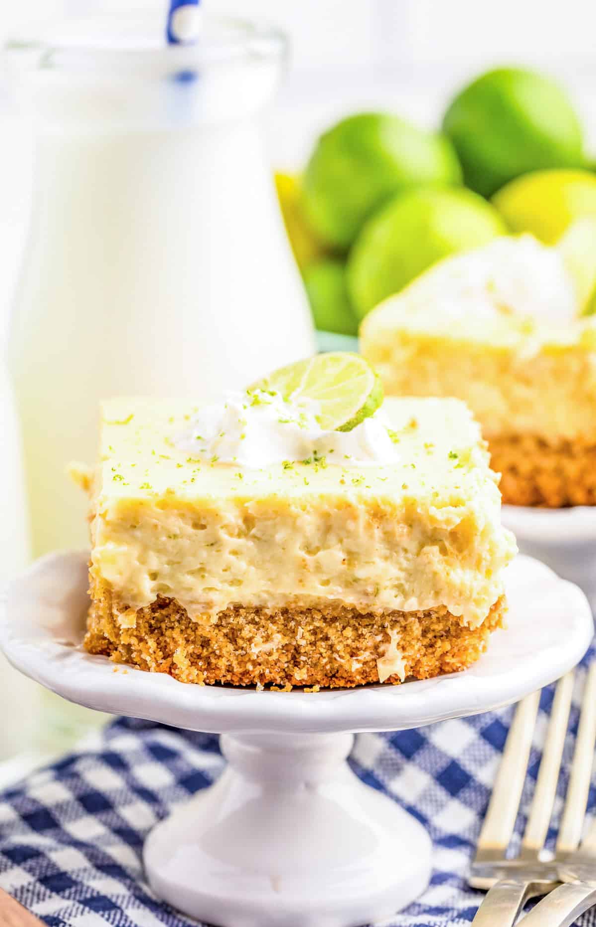 One of the Key Lime Pie Bars on mini white stand garnished with whipped cream, lime zest and lime slices with milk and limes in background.