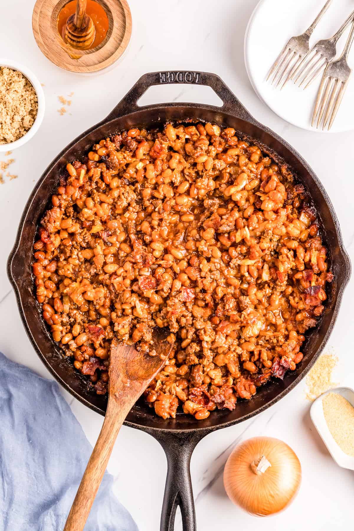 Overhead finished Baked Beans with Ground beef in cast iron skillet with wooden spoon and ingredients surrounding skillet.