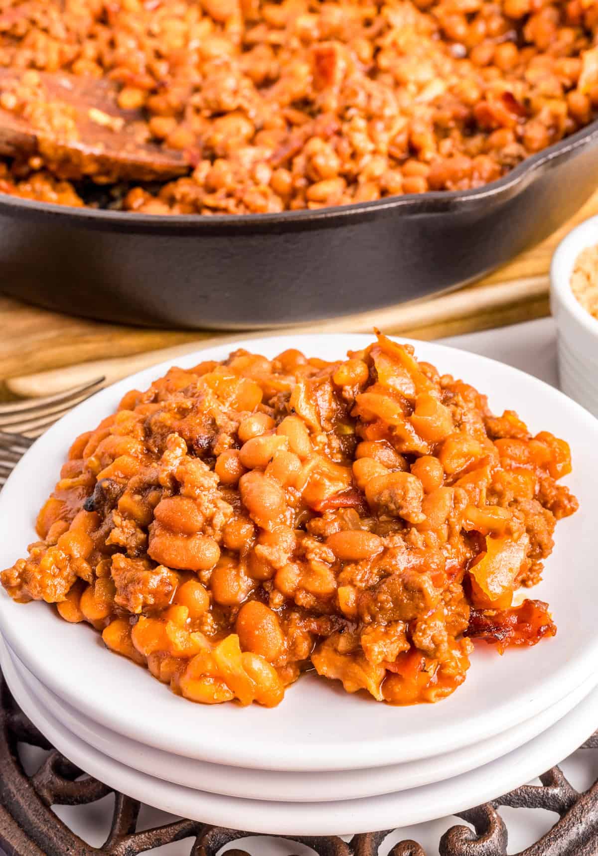 Baked Beans with Ground Beef on stacked white plate with skillet of beans in background.