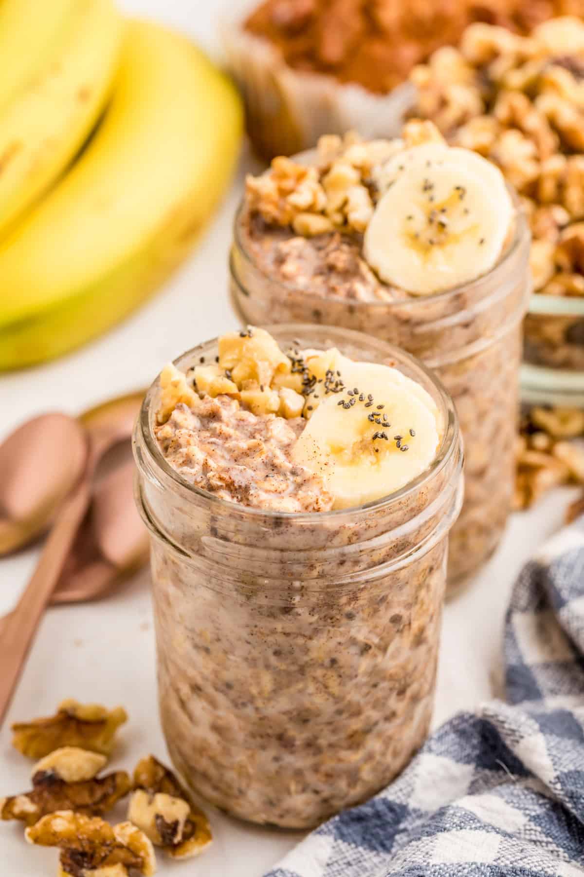 Two jars of Banana Overnight Oats topped with bananas, chia seeds and walnuts with other ingredients around jars.