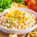 Close up square image of plated dip in white bowl with a chip in dip. Topped with shredded cheese and chopped cilantro.