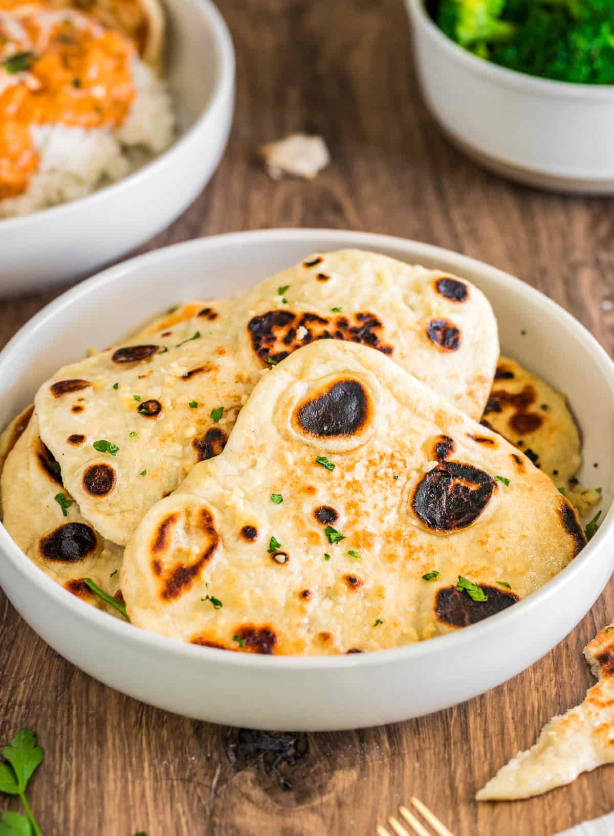 Garlic Naan in white bowl stacked on top of one another with butter chicken and herbs in background.