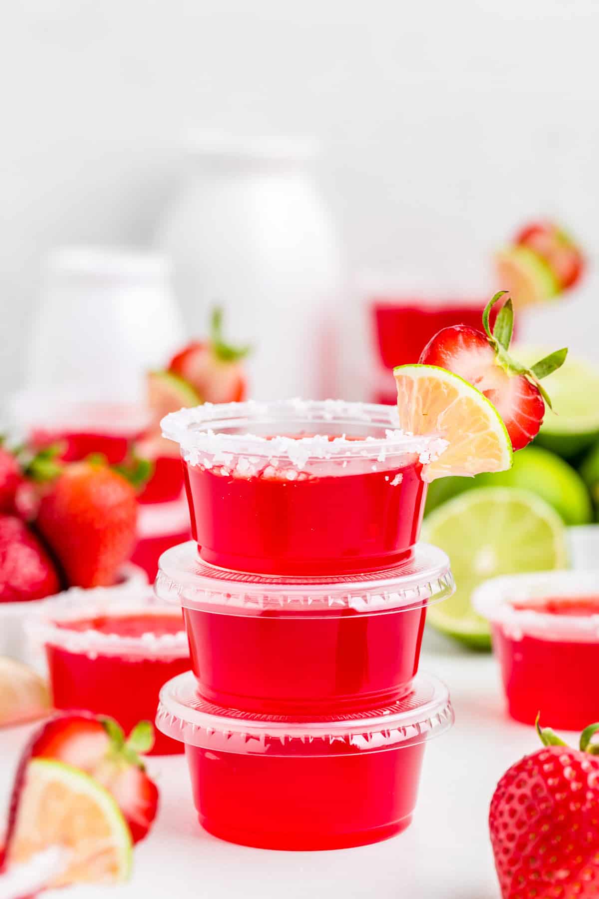 Three stacked Strawberry Margarita Jello Shots with top shot garnished with salt, lime and strawberry with other shots and ingredients around them.