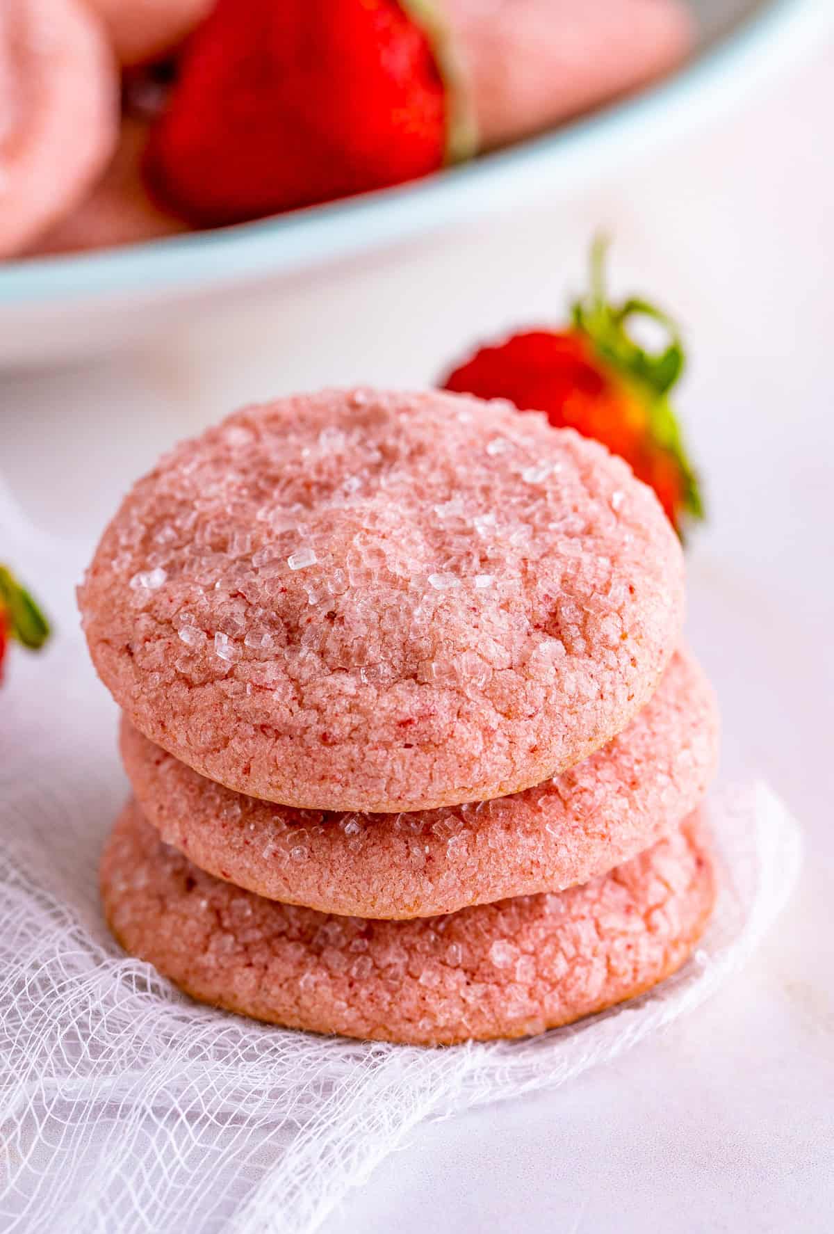 Three stacked Strawberry Sugar Cookies on white linen with strawberry behind them.