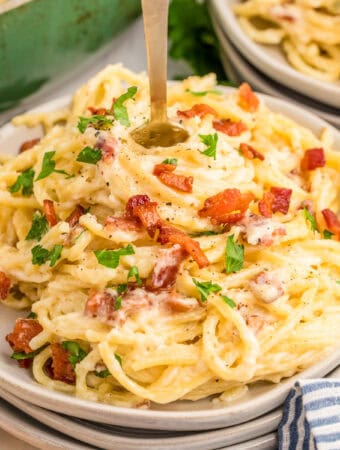 Square image of pasta on plate garnished with bacon and parsley with fork sticking out from center.