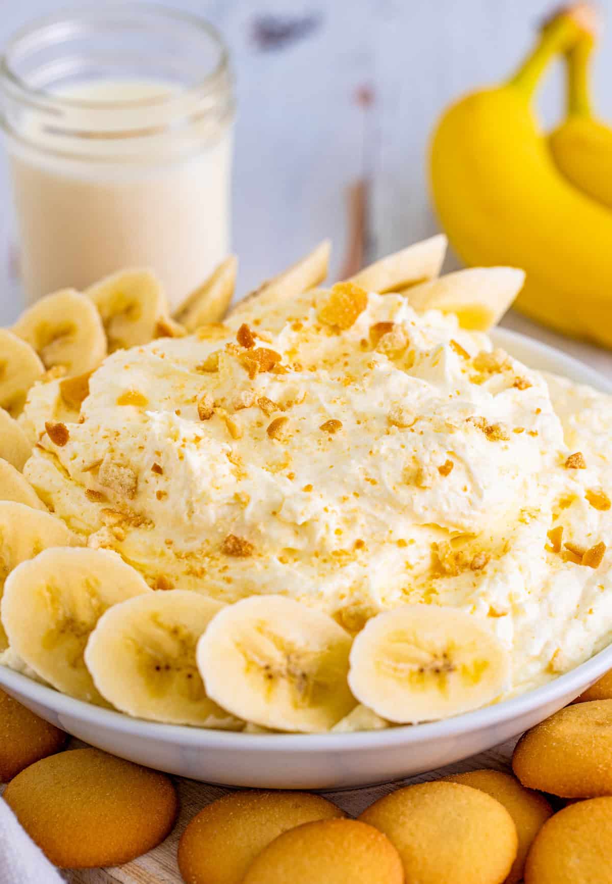 Banana Cream Pie Dip in white bowl with bananas and crushed vanilla wafers with milk and bananas behind it.
