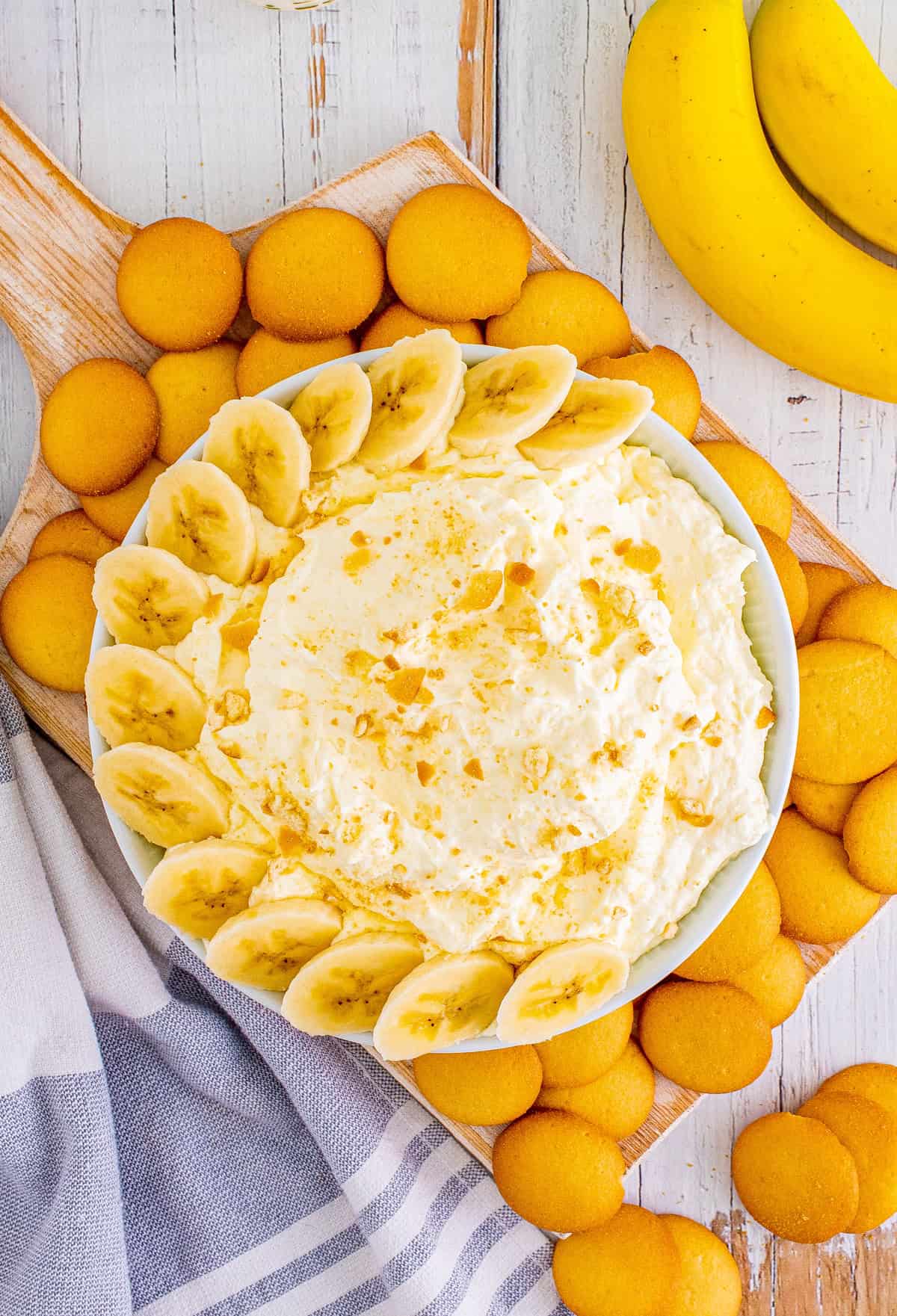 Overhead of Banana Cream Pie Dip in bowl surrounded by vanilla wafers and topped with garnishes of bananas and crushed vanilla wafers.