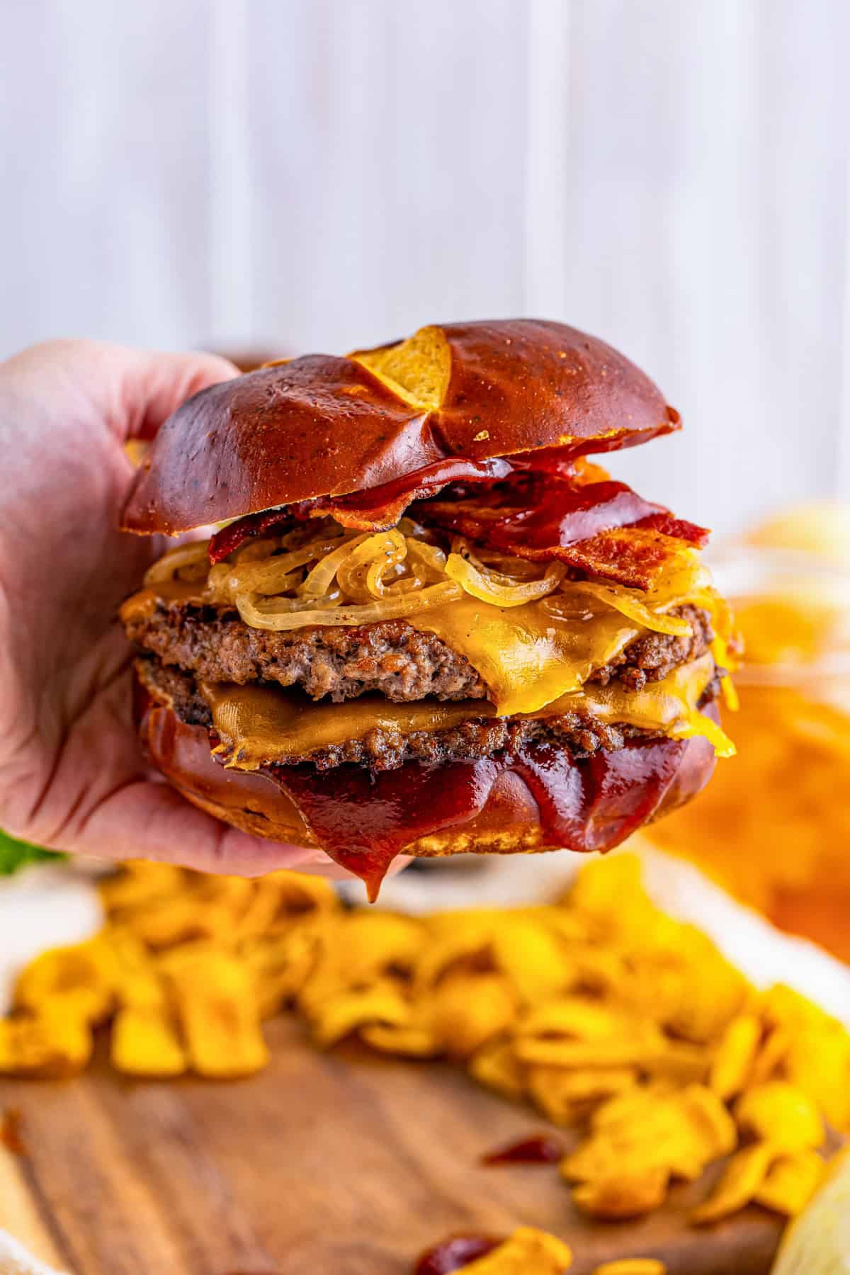 Hand holding up burger showing off all the layers with Fritos in the background on wooden board.