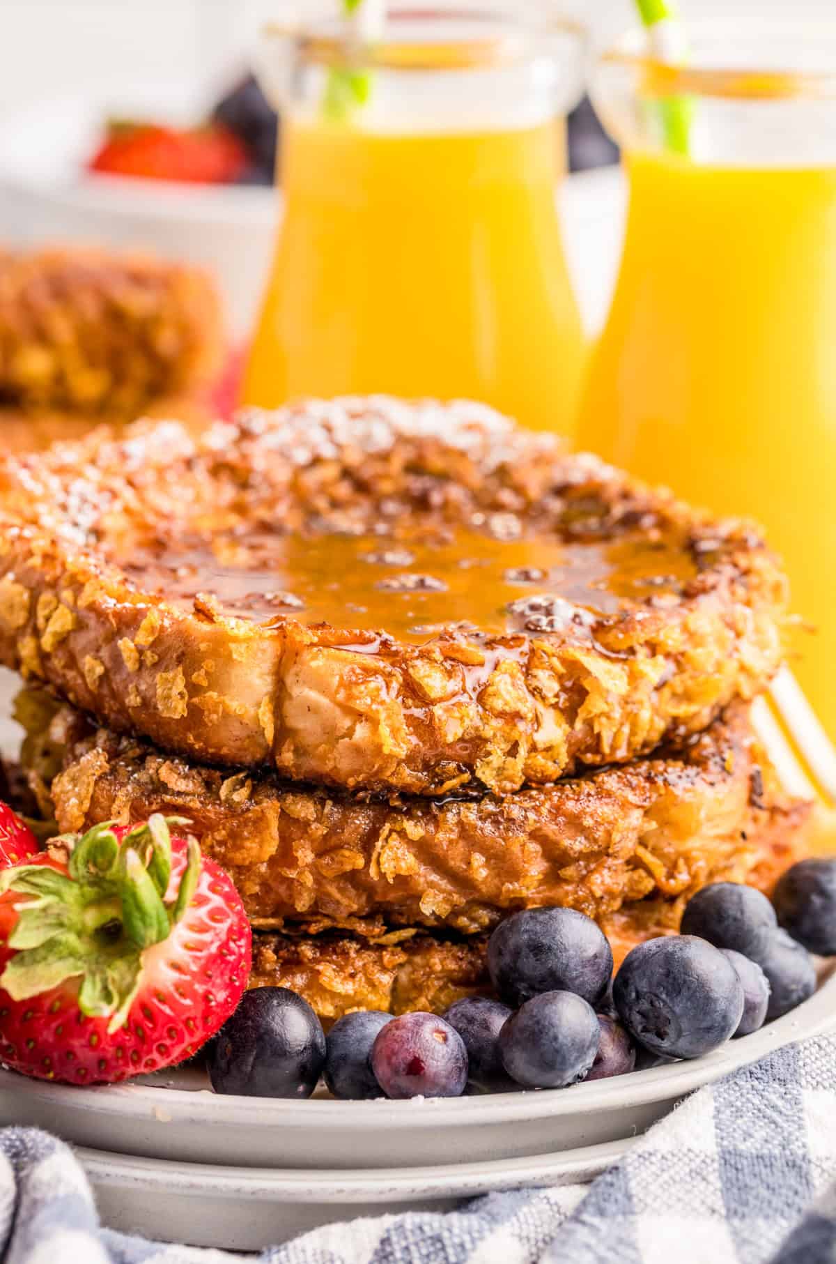 Stacked Crunchy French Toast on white plate with syrup and fruit and orange juice in background.