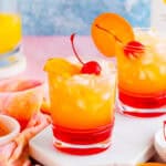 Square image of finished drink on white marble board with orange and cherry garnishes.