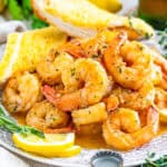 Close up square image of shrimp on white plate stacked on top of one another with lemons and garlic bread.