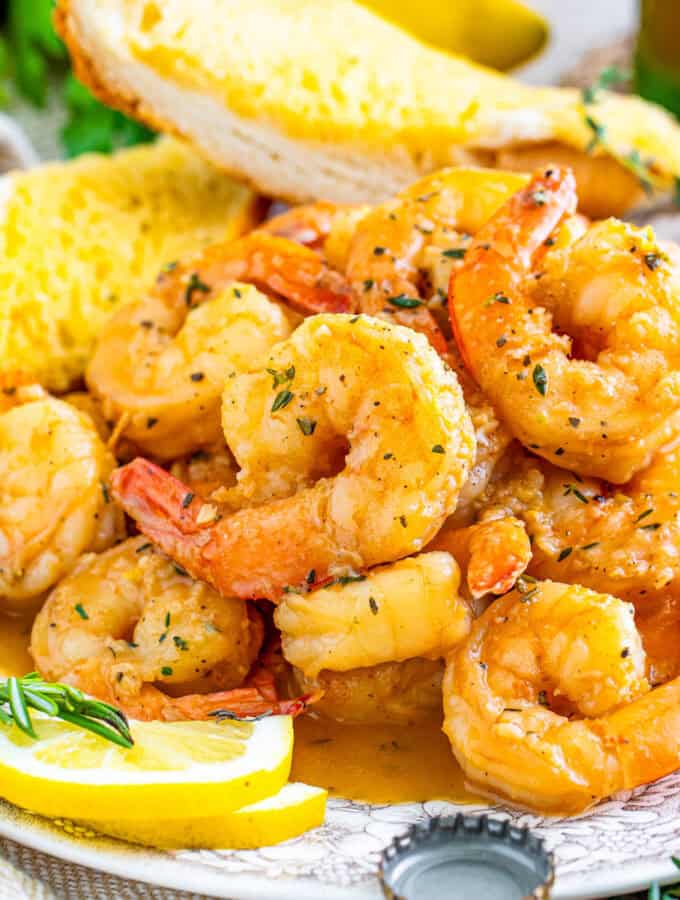 Close up square image of shrimp on white plate stacked on top of one another with lemons and garlic bread.
