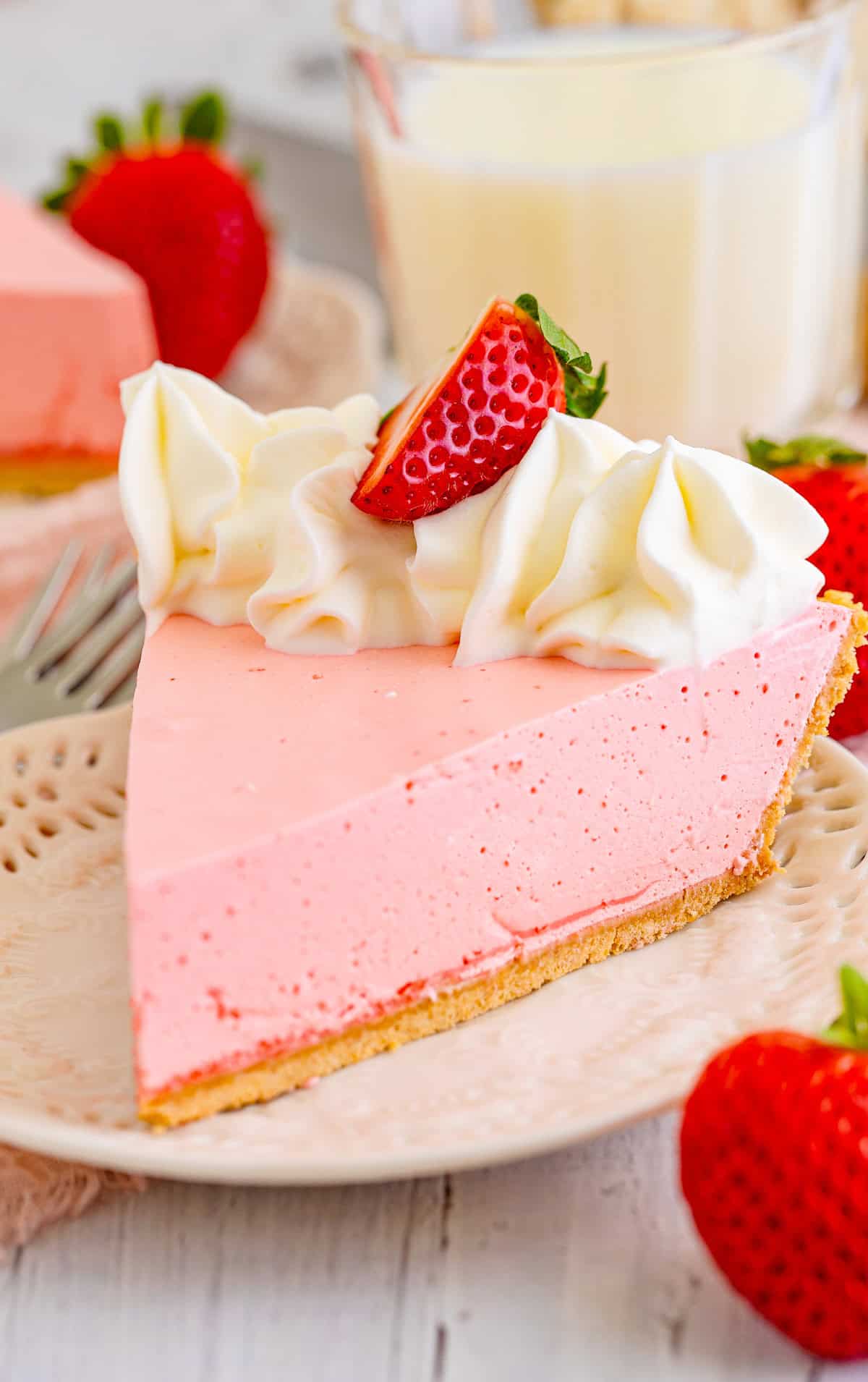 A slice of Strawberry Jello Pie on white plate with whipped topping and sliced strawberry with milk and strawberries around plate.