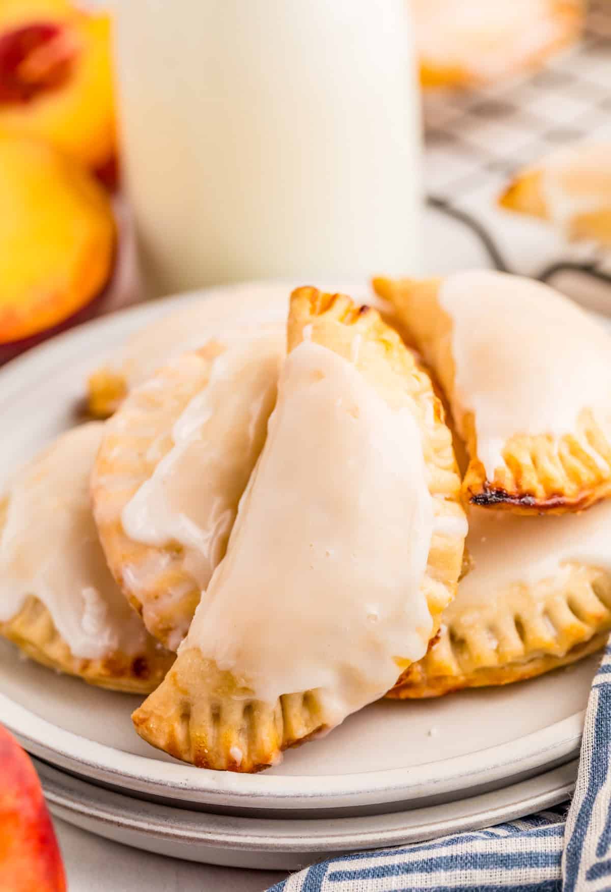 Peach Hand Pies stacked on white plate with glaze with milk and peaches in background.