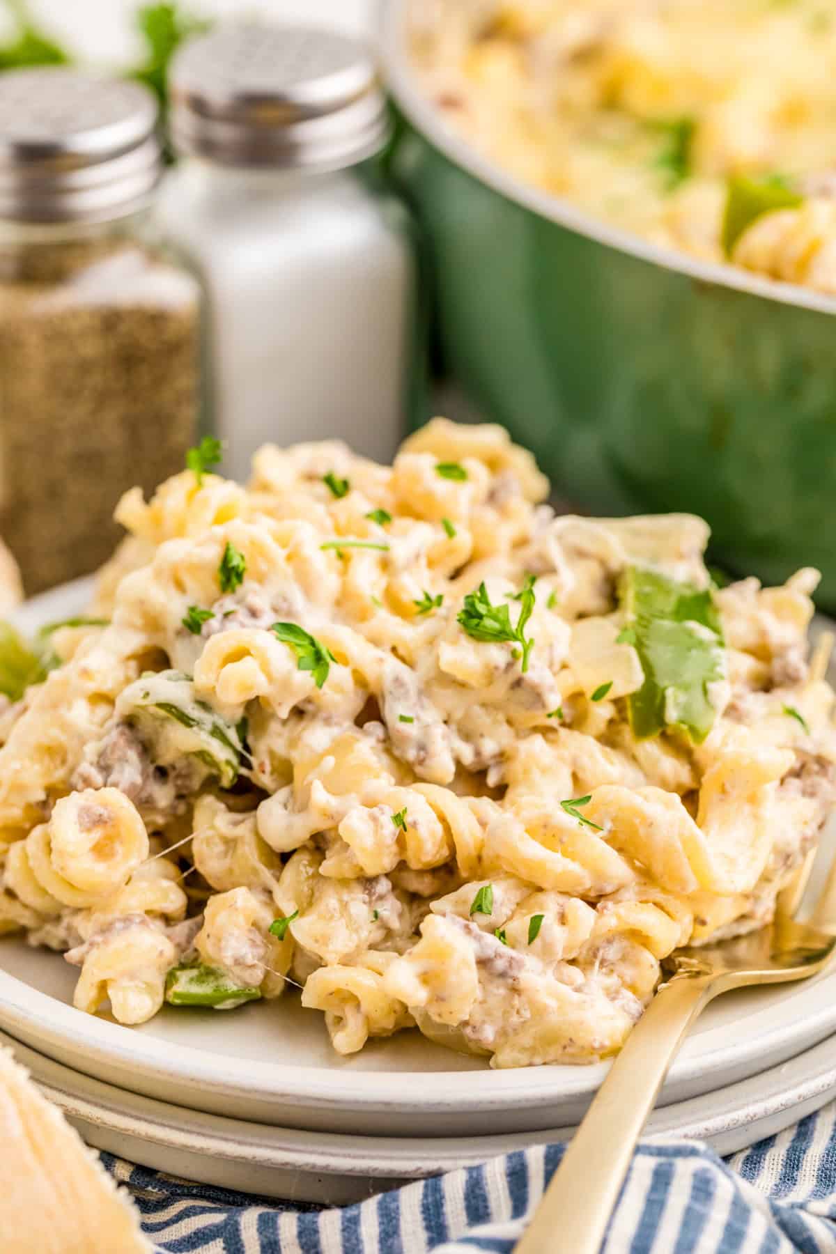 Philly Cheesesteak Pasta on white plate with fork with pan of pasta and salt and pepper in background.