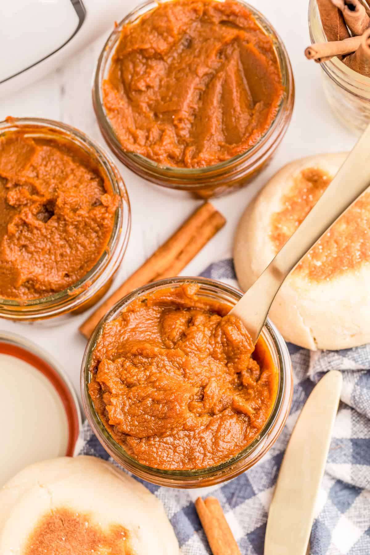 Overhead of Slow Cooker Pumpkin Butter in glass jars with a butter knife, cinnamon sticks and english muffins around the jars.