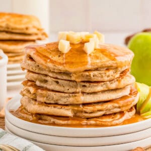 Close up square image of stacked pancakes on stacked plates with syrup and diced apples.