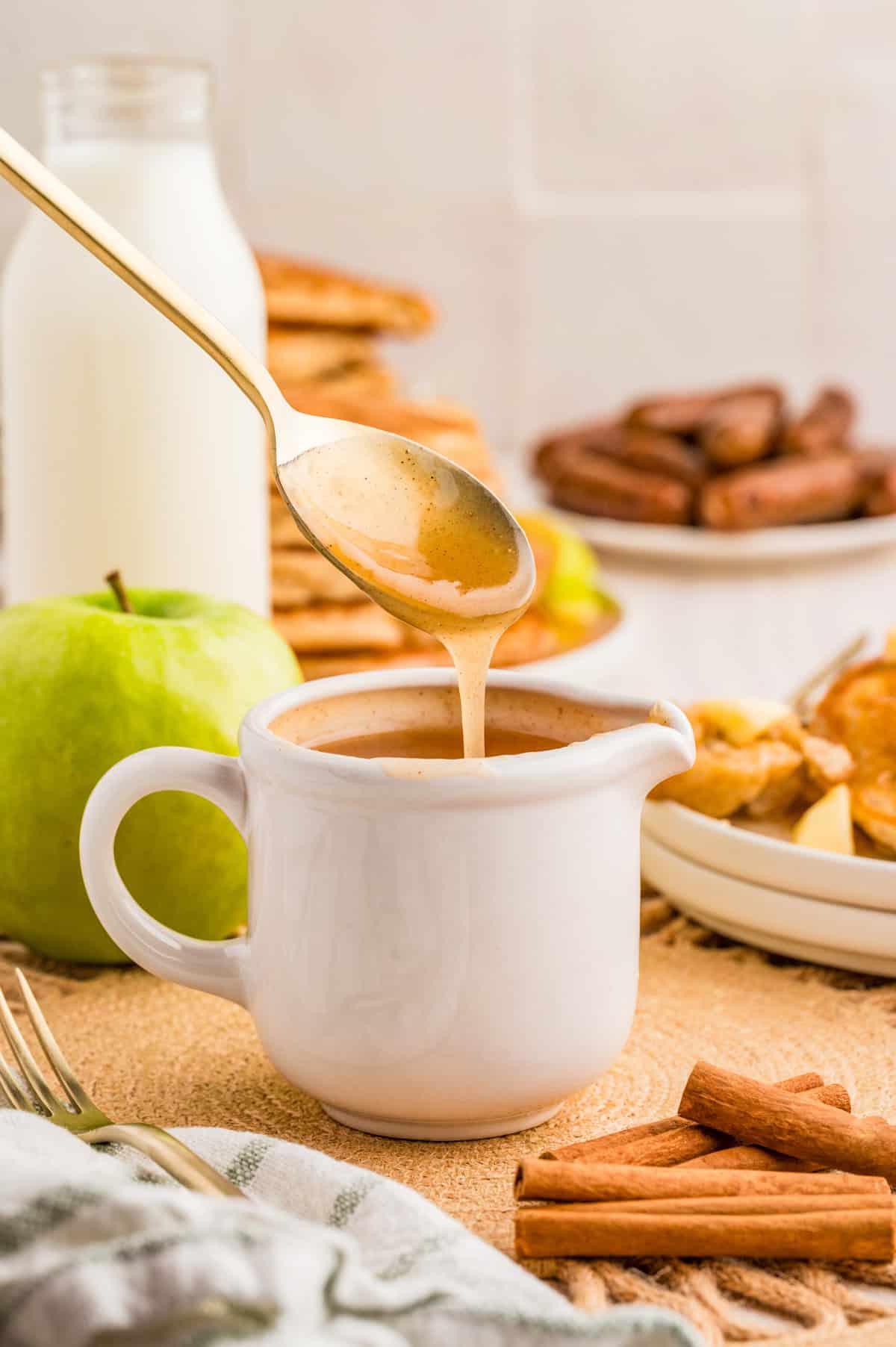 Apple Syrup in white spouted cup with spoon drizzling syrup into cup with apples and cinnamon in back and foreground.