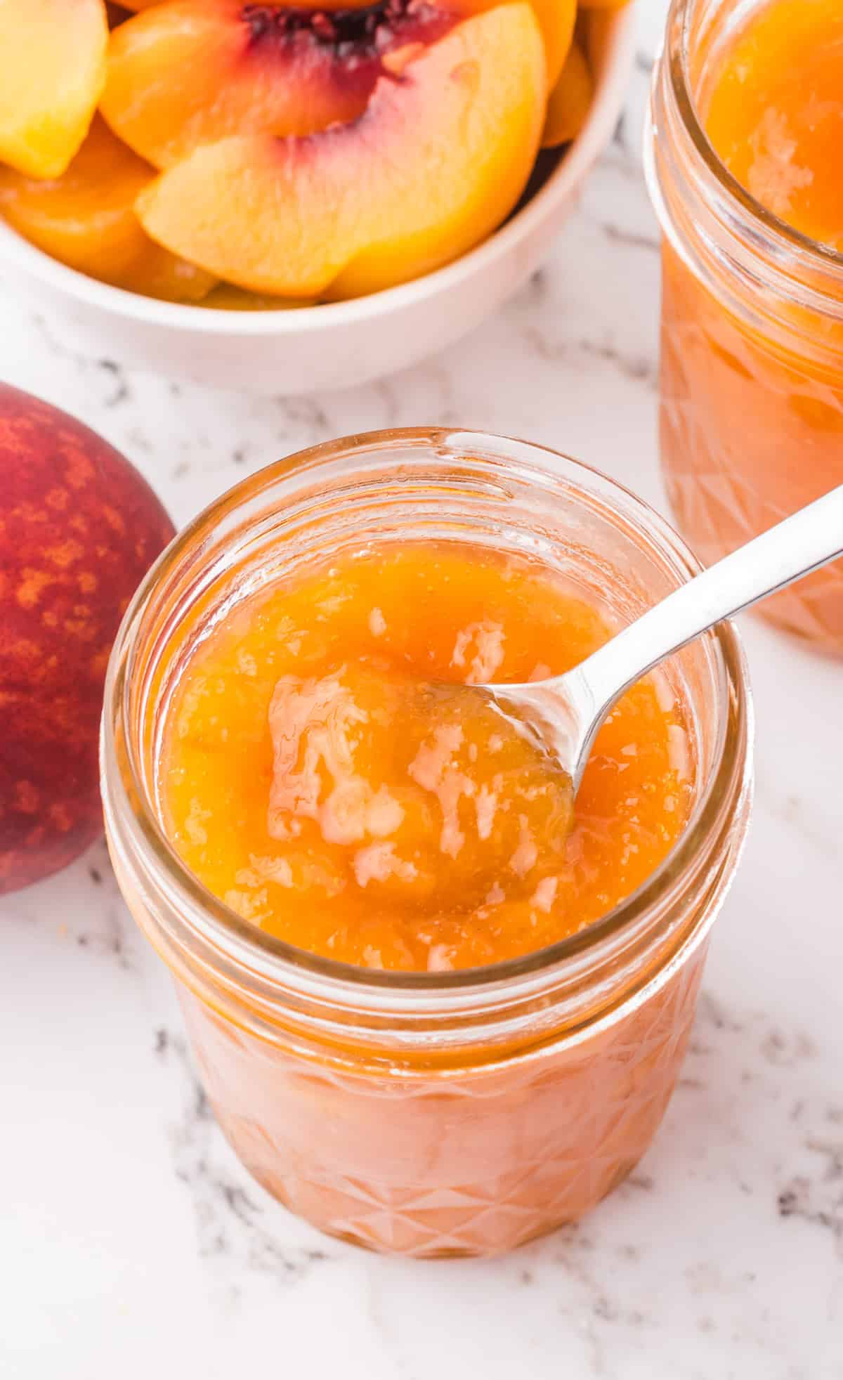 Overhead of jam in mason jar with a spoon in jar and peaches in bowl in background.