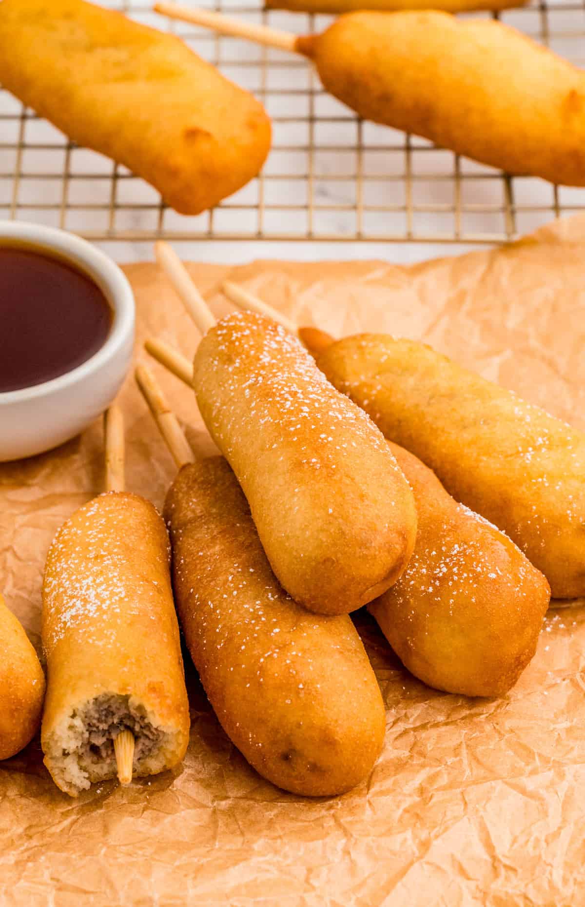 Stacked Breakfast Corndogs on parchment paper with bite taken out of one and syrup in background.