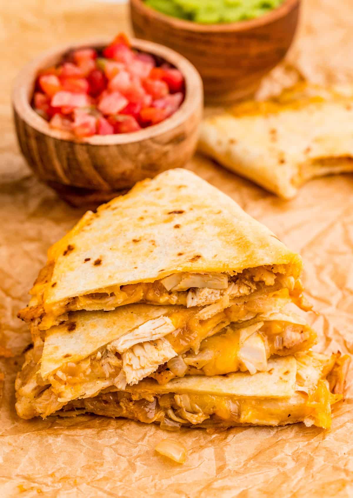 Chicken Quesadilla Recipe cut into triangles and stacked on parchment paper with tomatoes and guacamole in background.