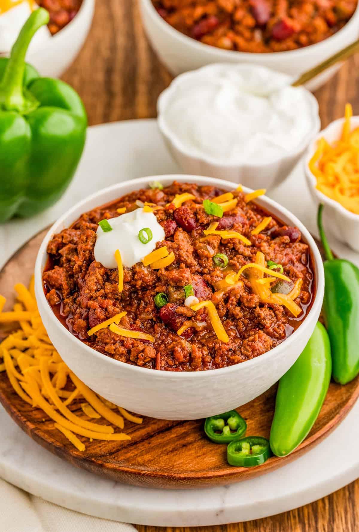 Chili Con Carne Recipe in white bowl on wooden plate topped with optional toppings.