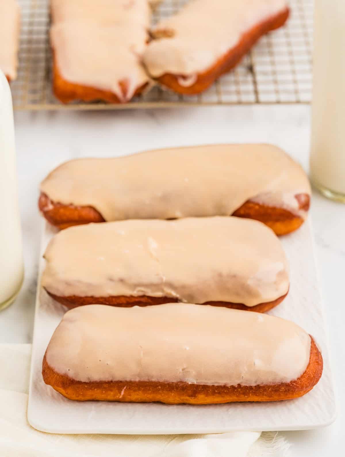 Three Maple Bars on white plate with milk at sides and more donuts in back.