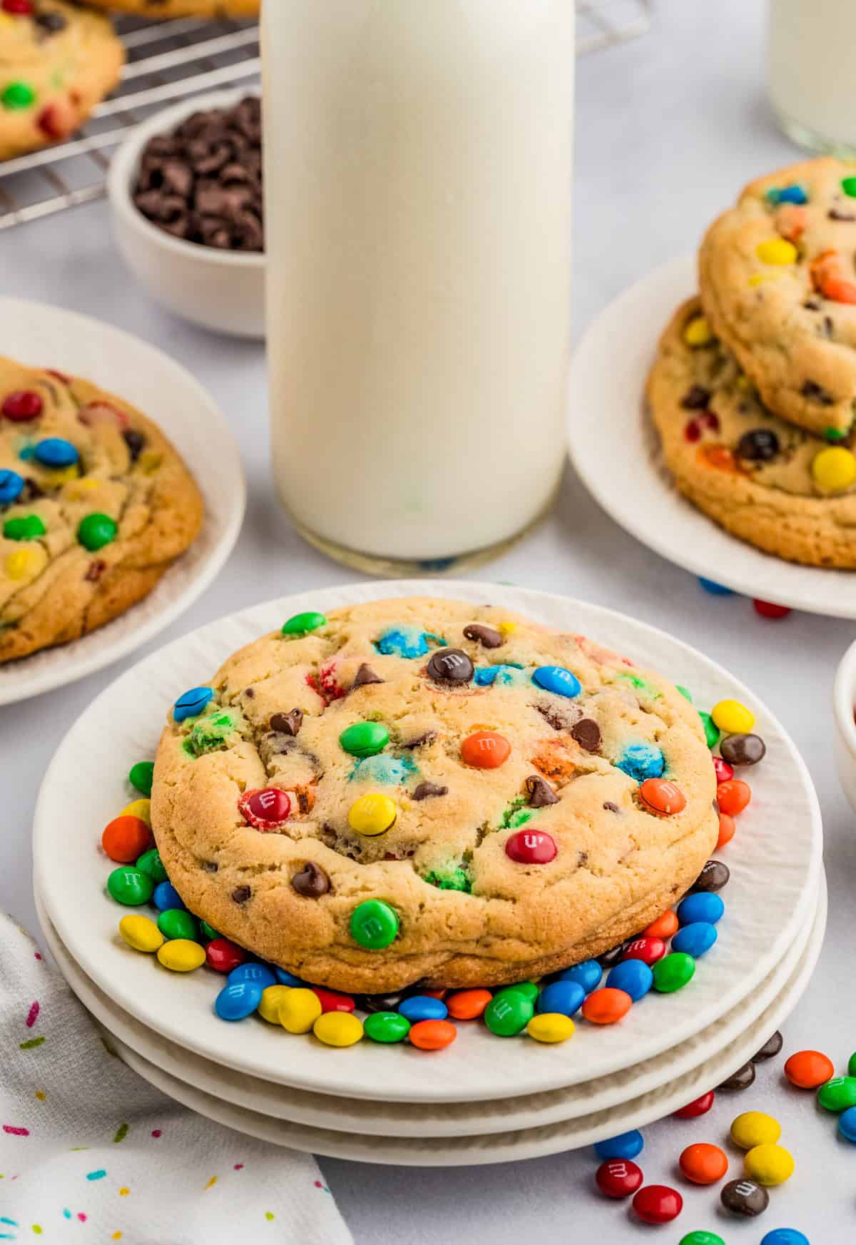 One of the M&M Chocolate Chip Cookies on white plates surrounded by mini M&M and milk in background.