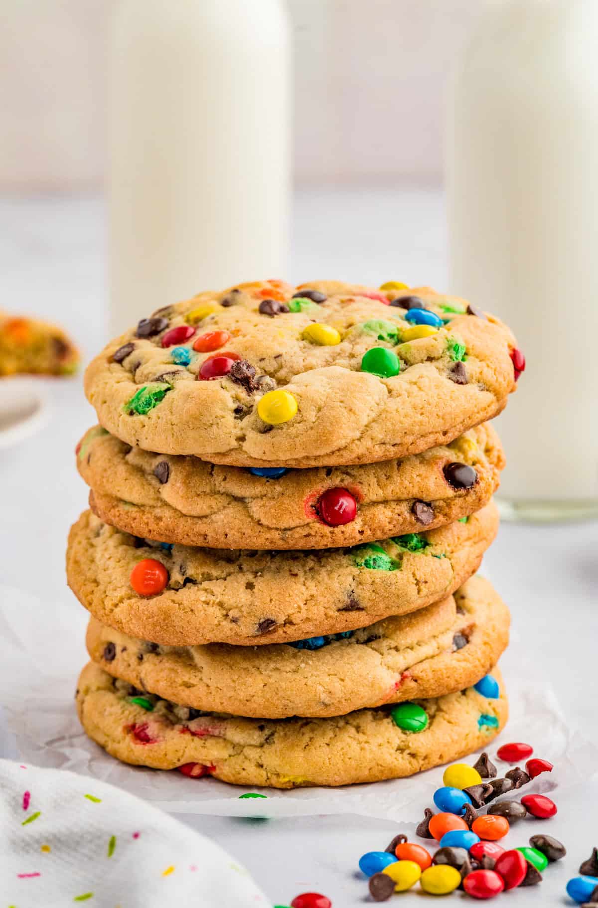 Five stacked M&M Chocolate Chip Cookies with mini M&M's and milk in background.