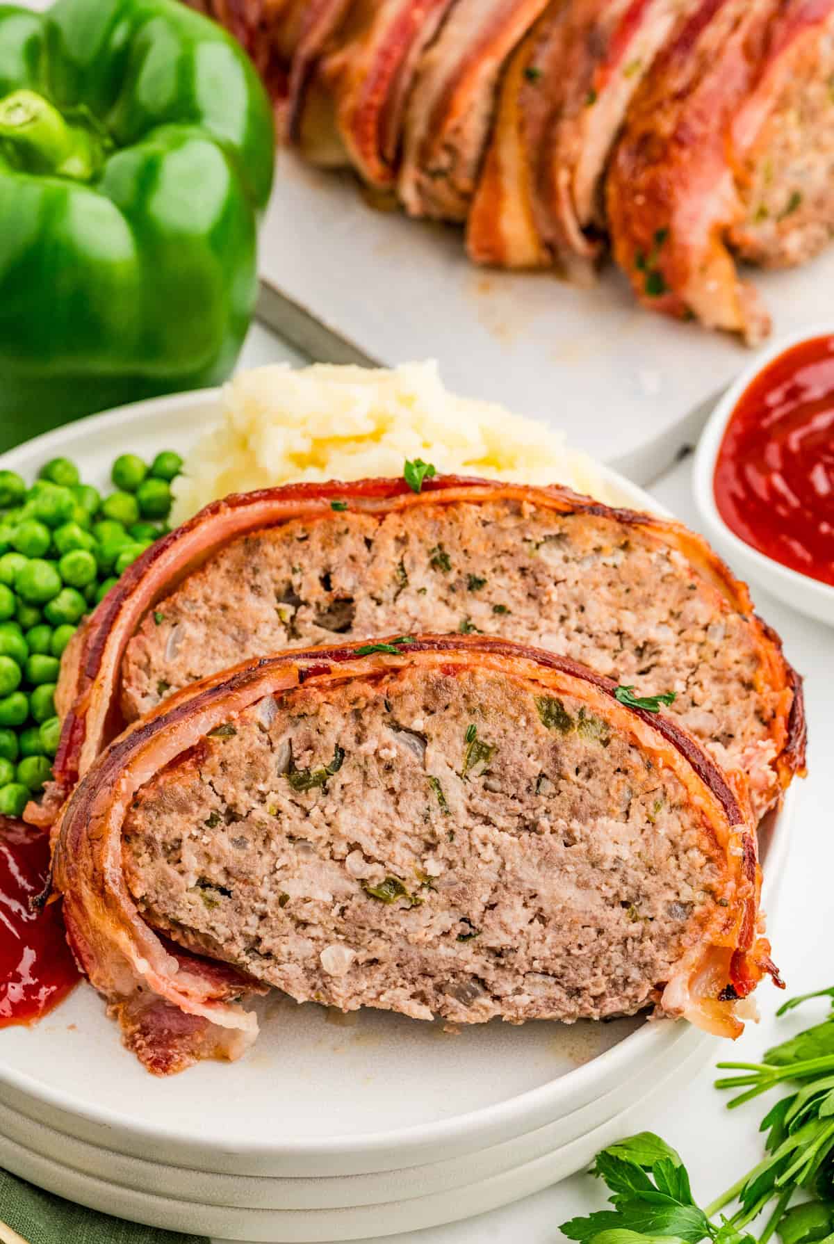 Two slices of Bacon Wrapped Meatloaf on plate with potatoes, peas and ketchup.