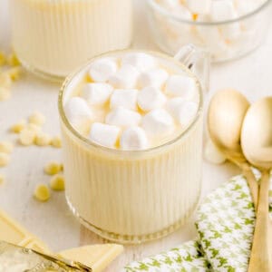 Square image of hot chocolate in glass with marshmallows white chocolate and gold spoons near glass.