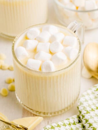 Square image of hot chocolate in glass with marshmallows white chocolate and gold spoons near glass.