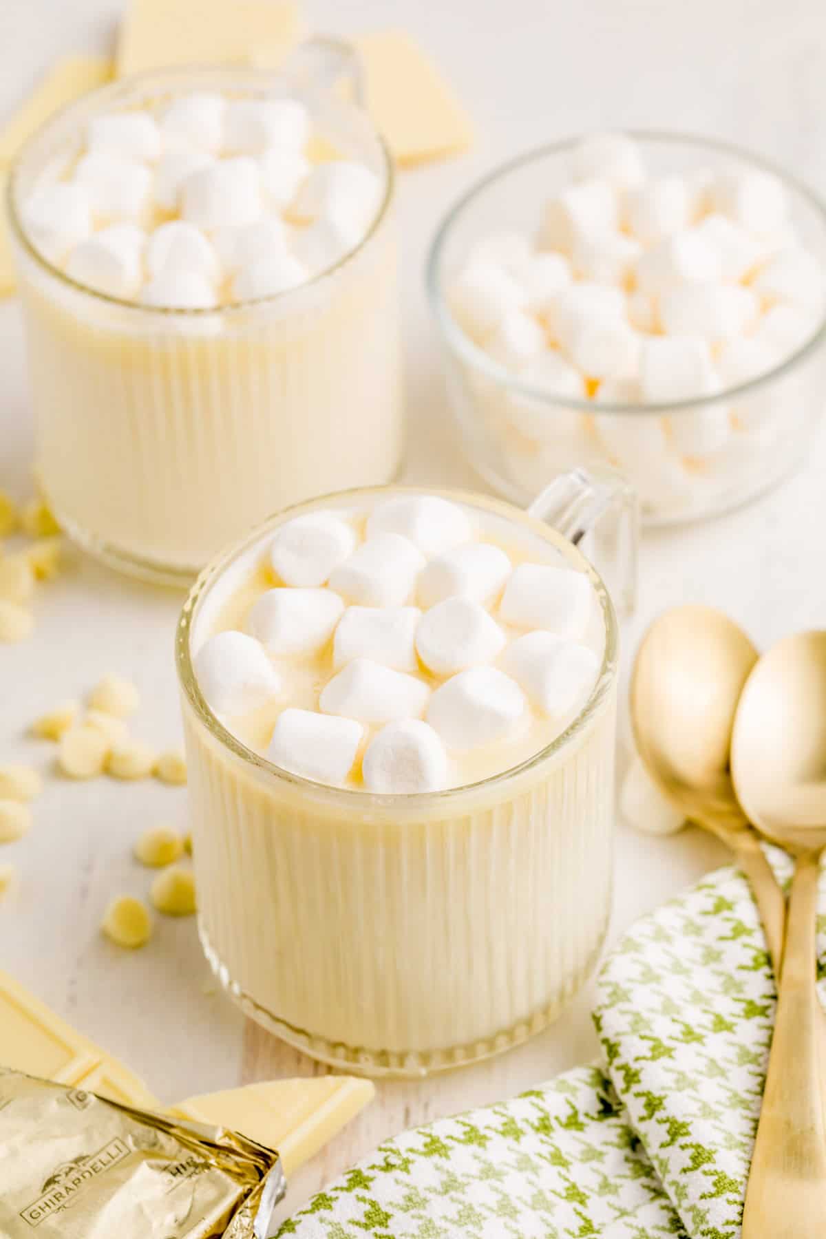 Easy White Hot Chocolate in ribbed clear glasses topped with marshmallows with white chocolate and gold spoons beside glasses.