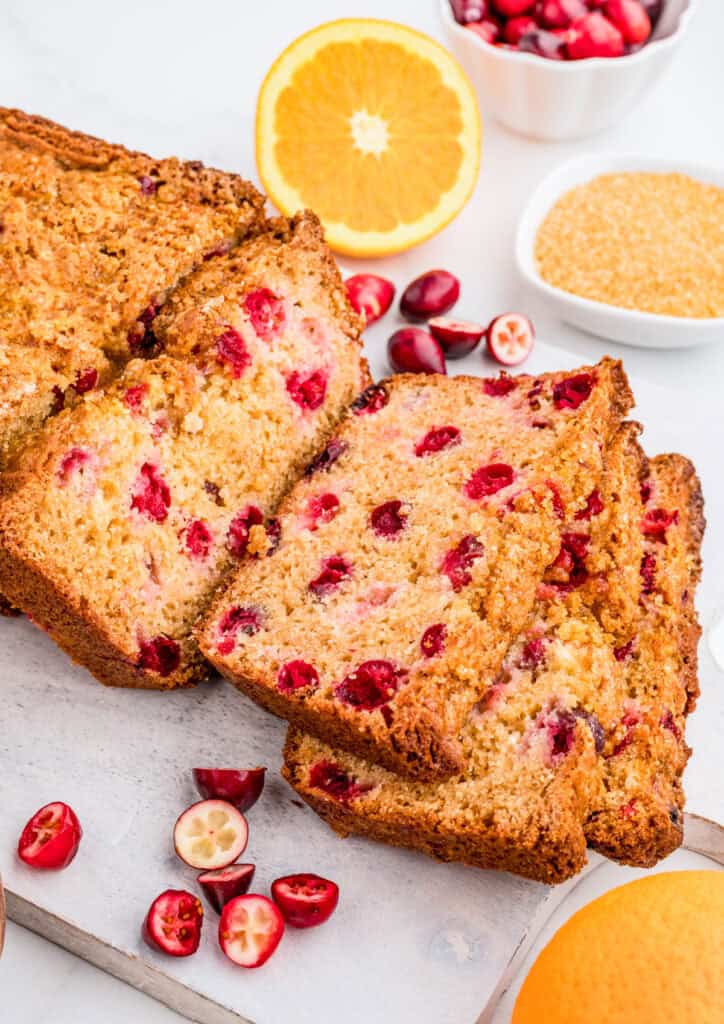 Cranberry Bread cut into slices on white board off of the whole loaf with cranberries and oranges around it.