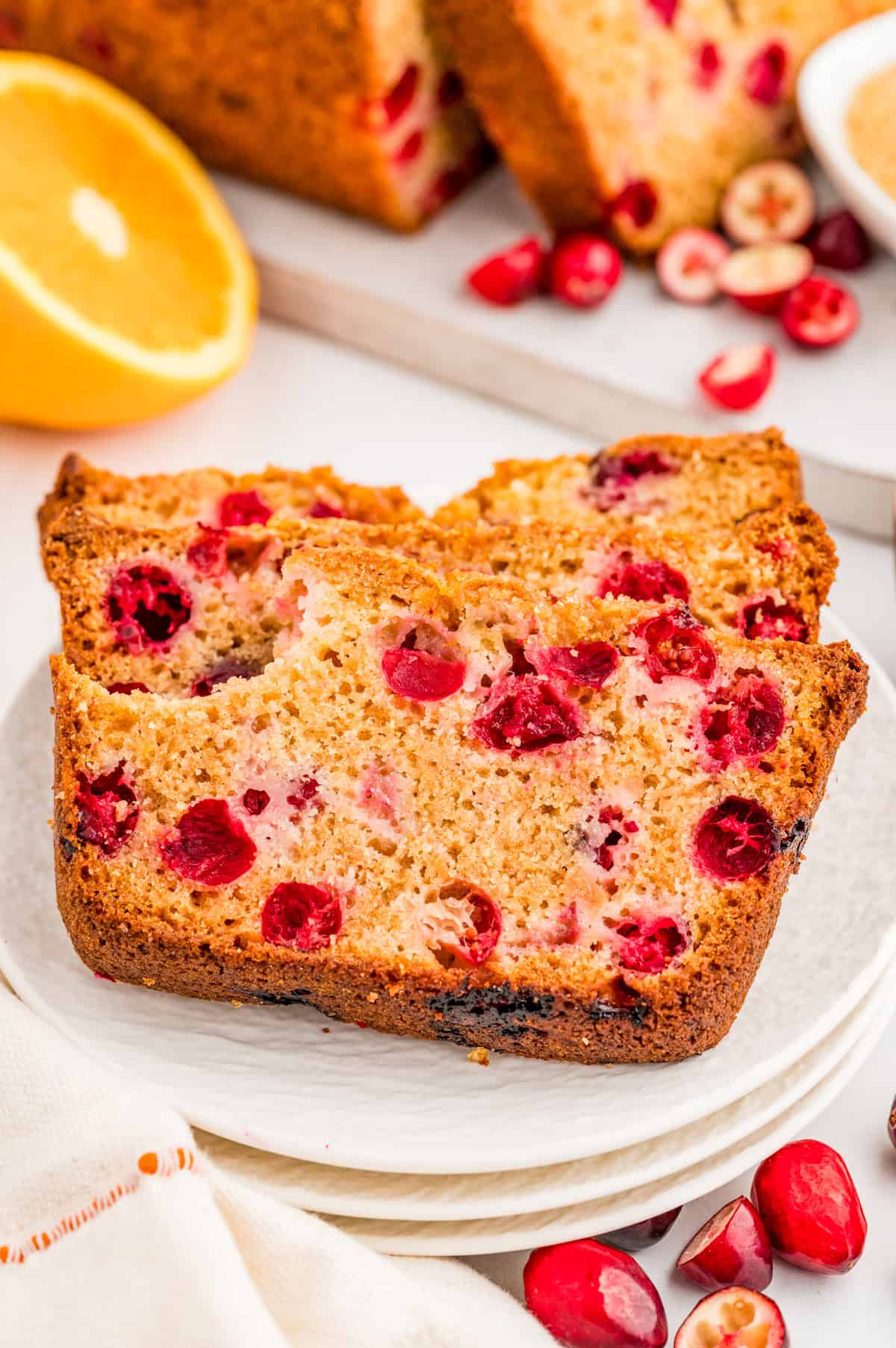 Two slices of Cranberry Bread on white plate with sliced orange in background and cranberries in front.