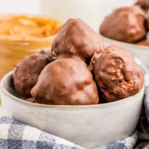 Square image of peanut butter balls in bowl with peanut butter in background.