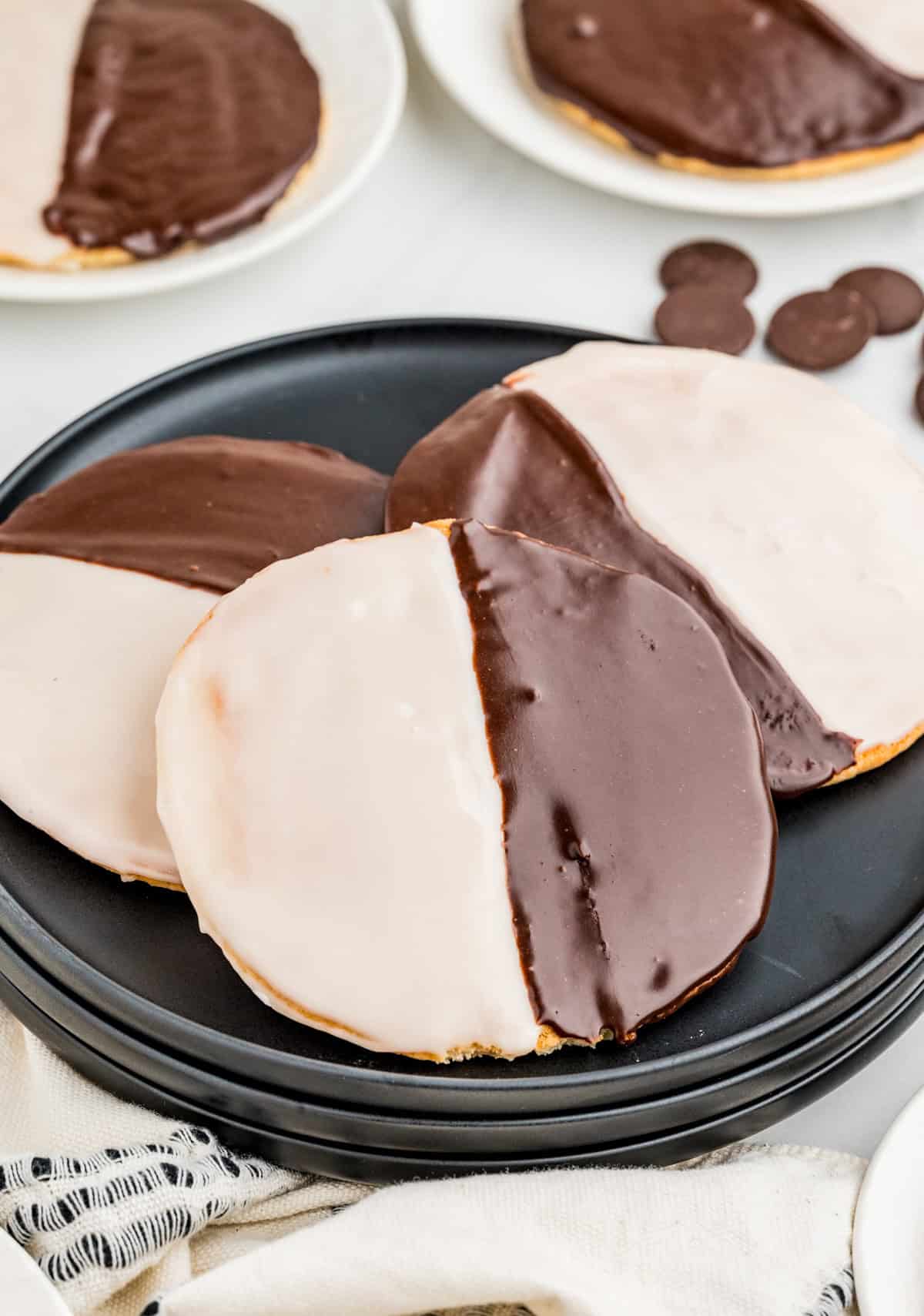 Three Black and White Cookies layered on black plate with more cookies in background.