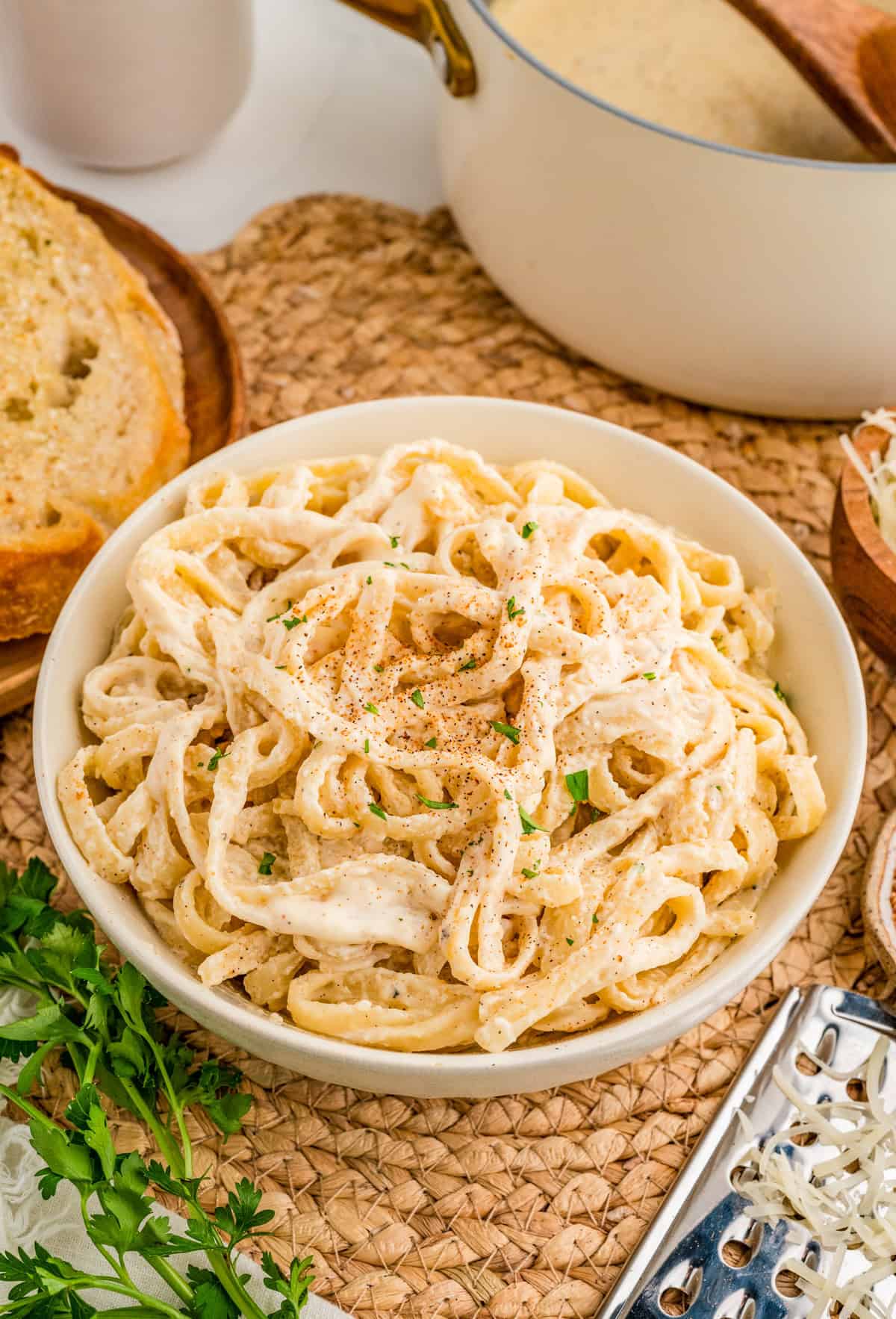 Cajun Alfredo Sauce over pasta in white bowl on wicker trivet with bread and parsley around bowl.