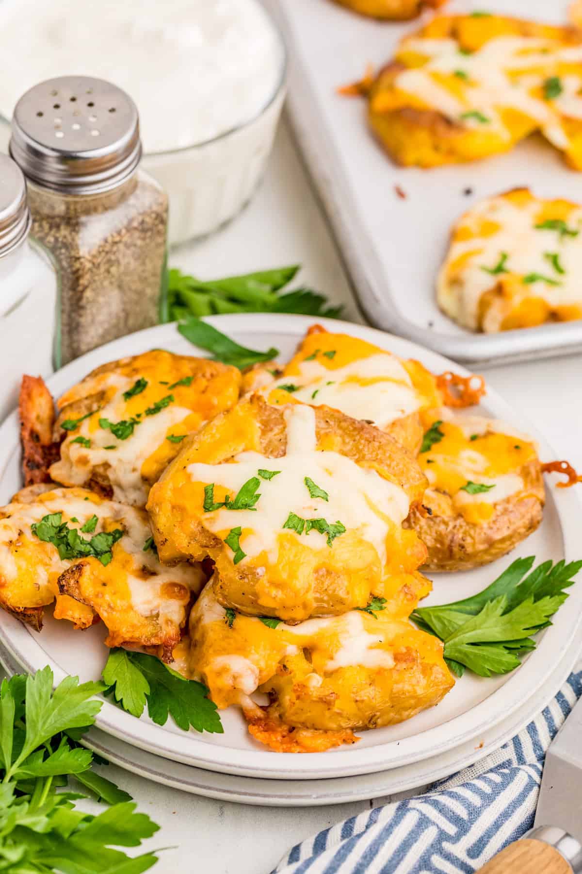 Cheesy Smashed Potatoes layered on white plate topped with parsley with more potatoes on baking sheet in background.