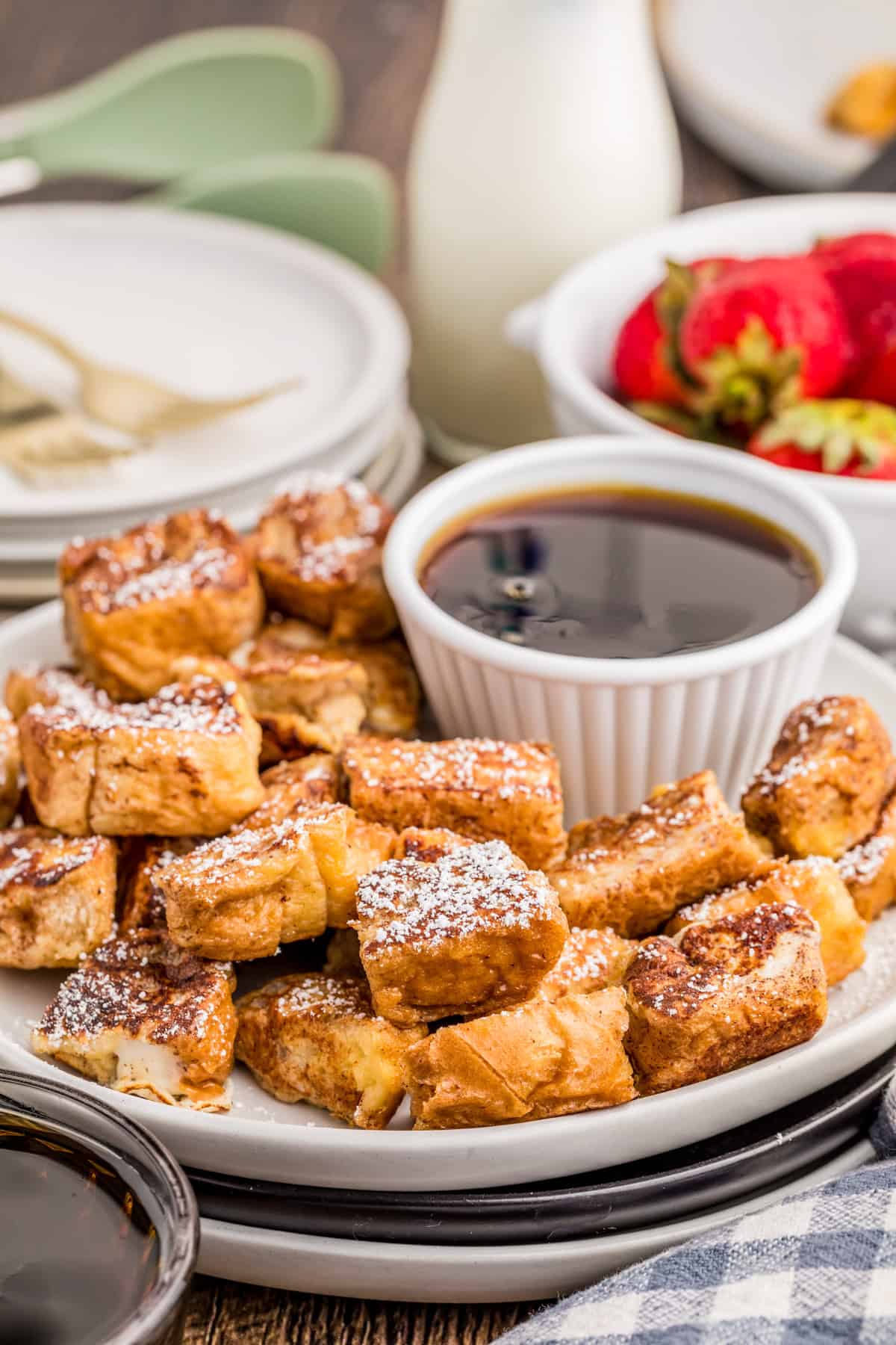 French Toast Bites on white plate topped with powdered sugar with a bowl of syrup on plate.