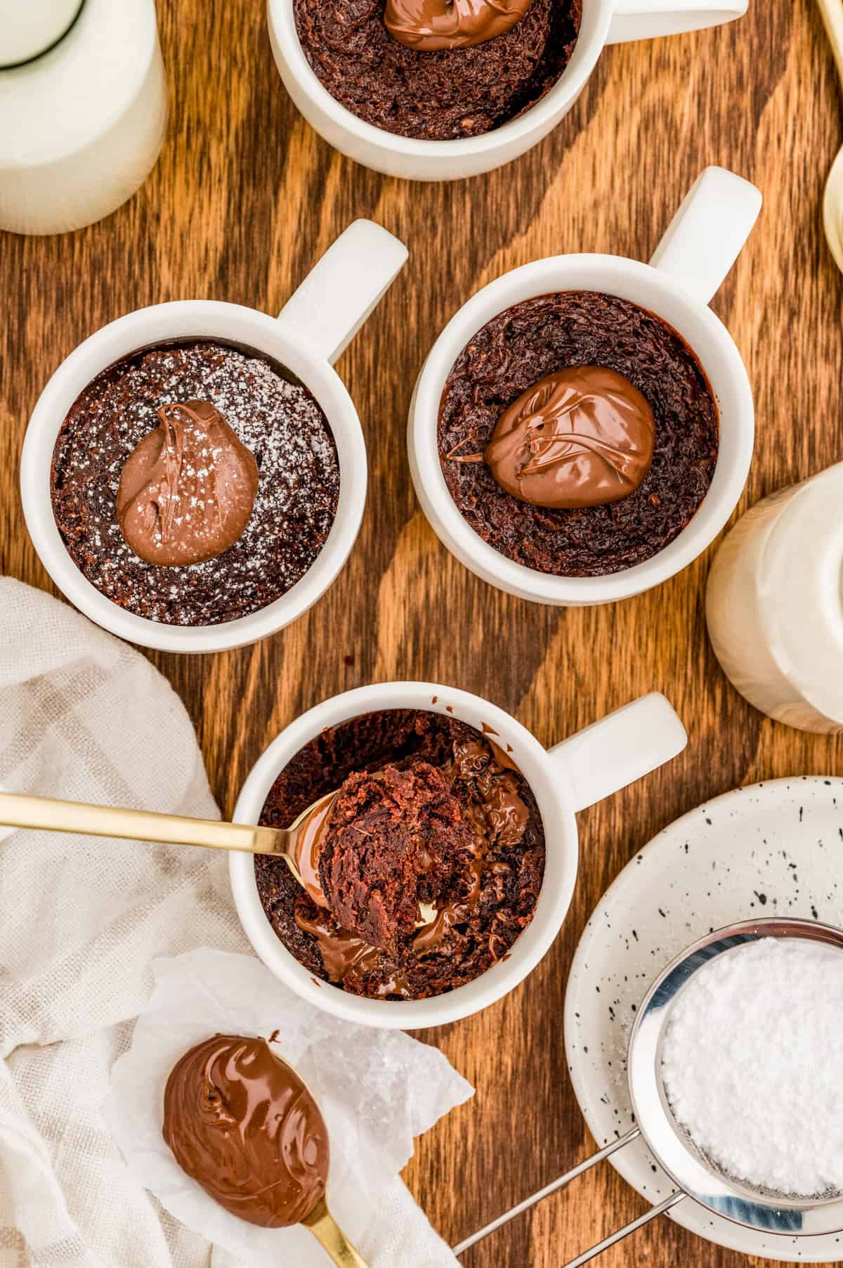 Overhead of finished cakes topped with nutella and powdered sugar with one mug having a spoon scoop out cake.