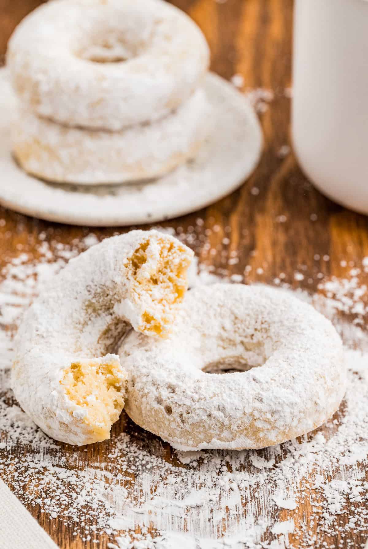 Powdered Sugar Donuts on a plate and wooden board with one donut with bite taken out of it.