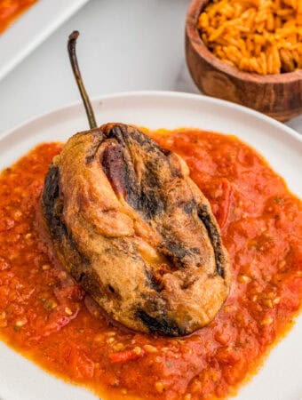 Square image of finished chile on white plate over sauce with mexican rice in background.