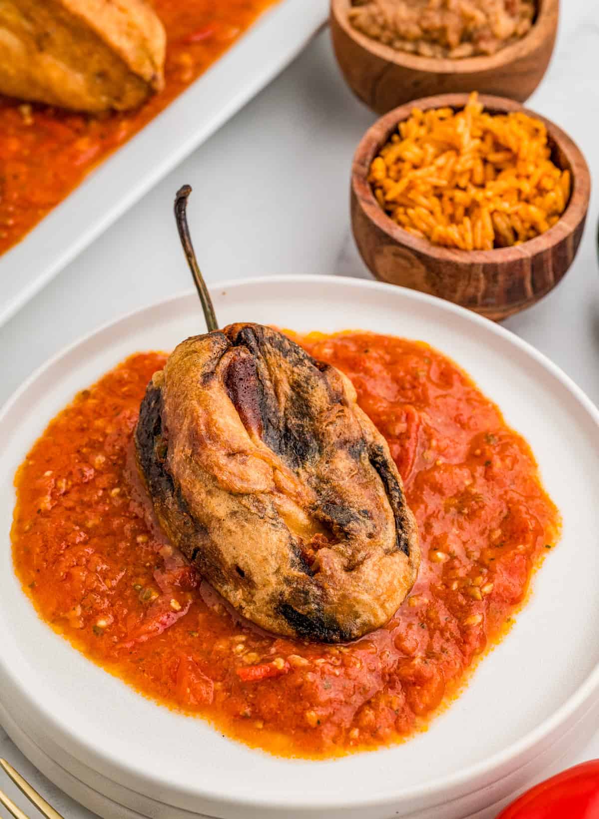 Finished Chile Rellenos over sauce on white plate with mexican rice and refried beans in background.